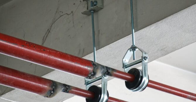 Two silver and black pipe supports hold two pipes that run near a room's ceiling in place. Both of the pipes are red.