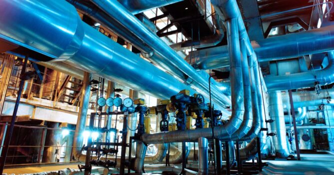 Many pipes run through an industrial setting, featuring gauges, valves, and ladders. The room has blue lighting.