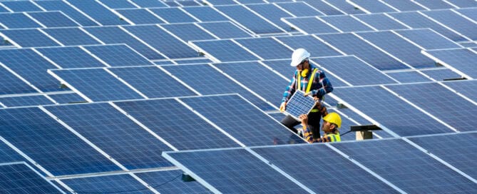 A pair of engineers work on the panels outside a solar power plant station. They stand in the middle of many rows of panels.