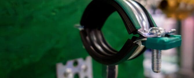 A green and black pipe anchor features a double-screw clamp. It stands on a screw against a green background.