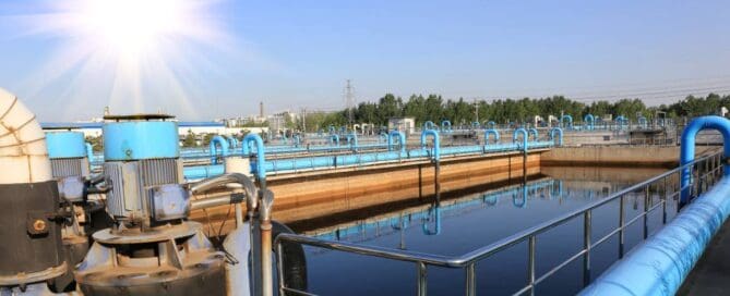 Large blue pipes run outside the exterior of a wastewater treatment plant. The sun shines brightly outside.