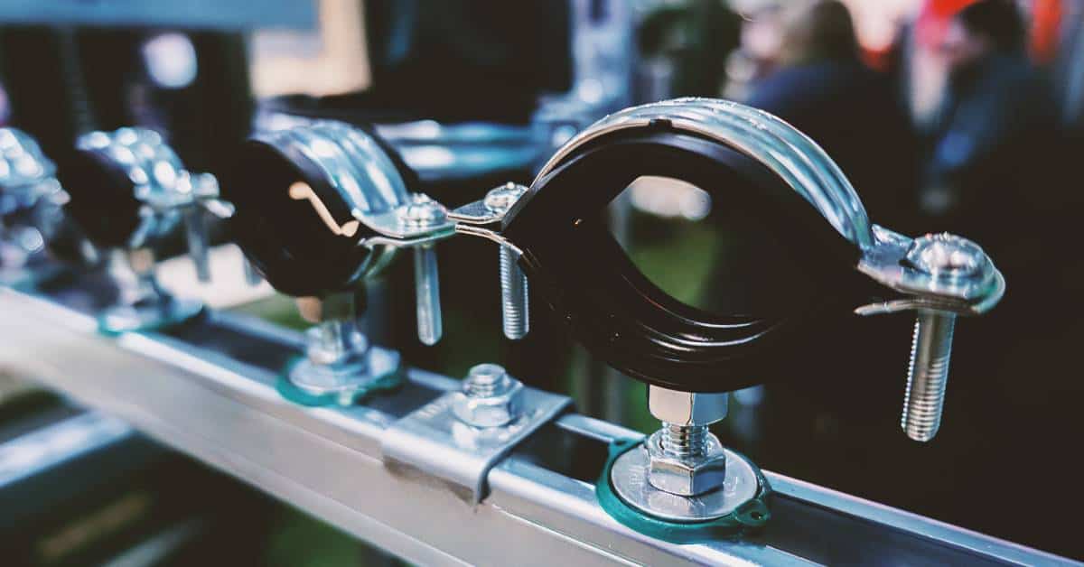 Several black and silver pipe clamps sit in a row on a metal beam. Two people sit in the background behind them.