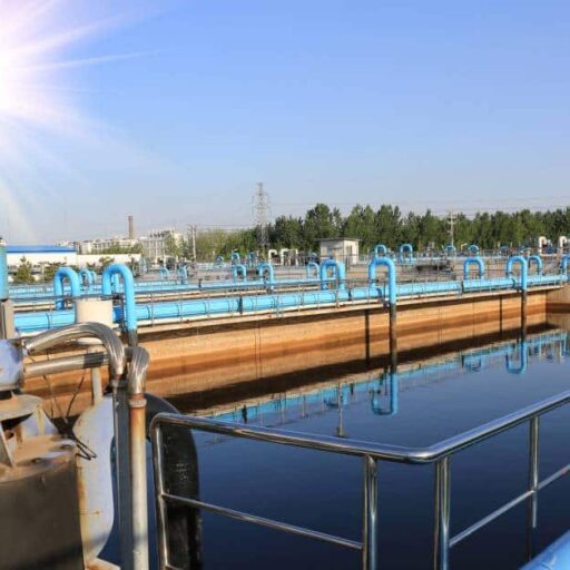 Large blue pipes run outside the exterior of a wastewater treatment plant. The sun shines brightly outside.