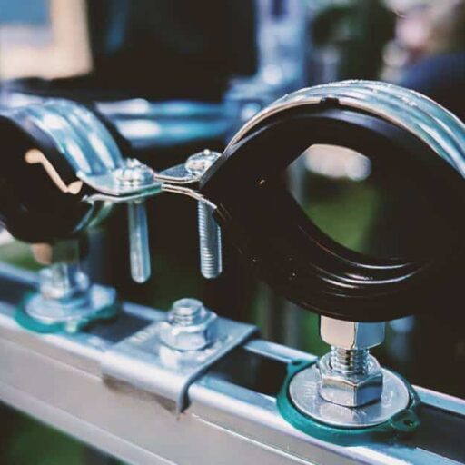 Several black and silver pipe clamps sit in a row on a metal beam. Two people sit in the background behind them.